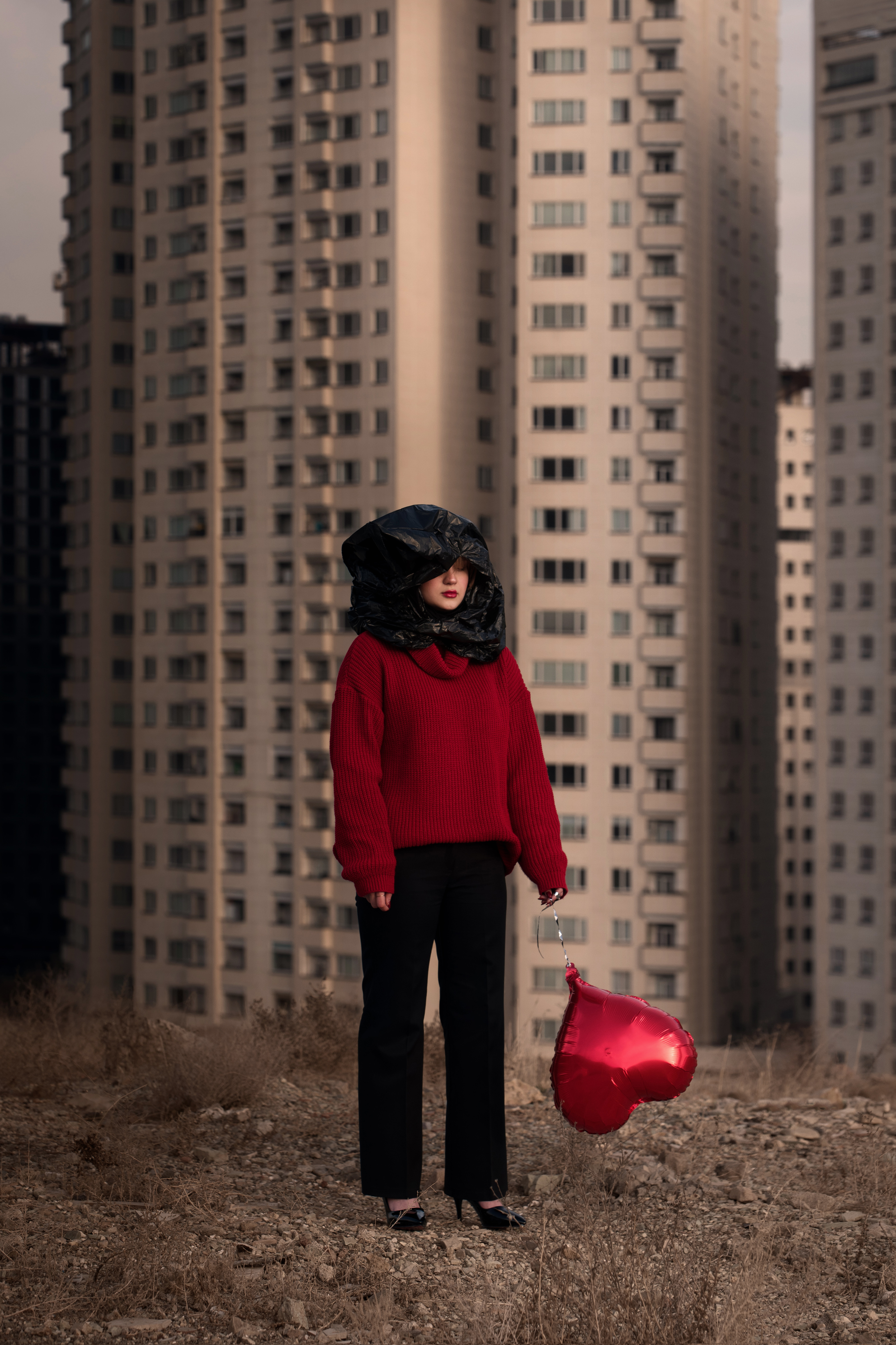 A woman stands in front of a block of flats. She's wearing a red sweater, a bin bag partly covering her face and a red balloon shaped like a heart.© Siavosh Ejlali, Iran, Islamic Republic Of, Winner, Open Competition, Creative, 2026 Sony World Photography Awards