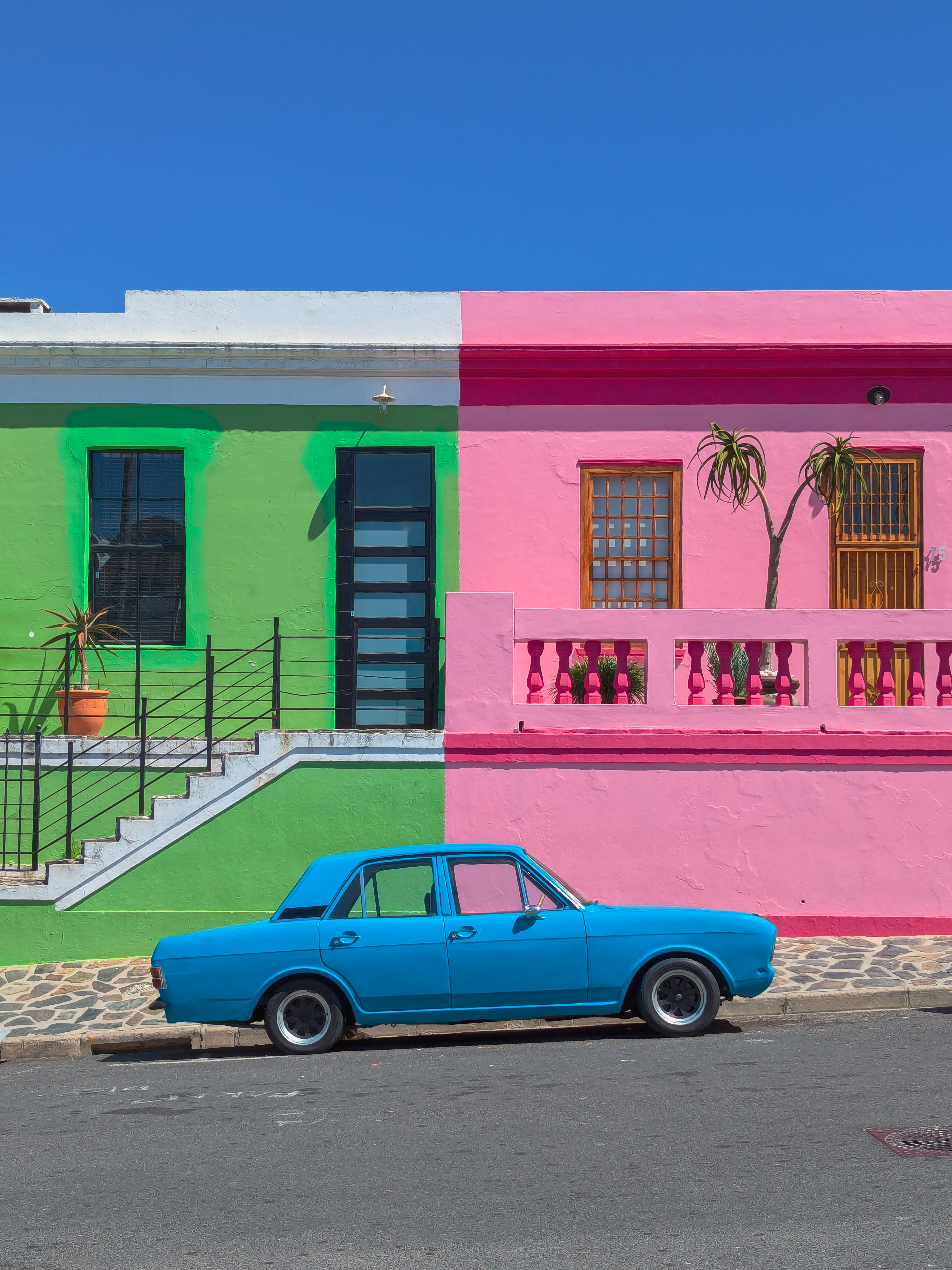 A blue, vintage car is parked in front of green and a bright pink buildings.