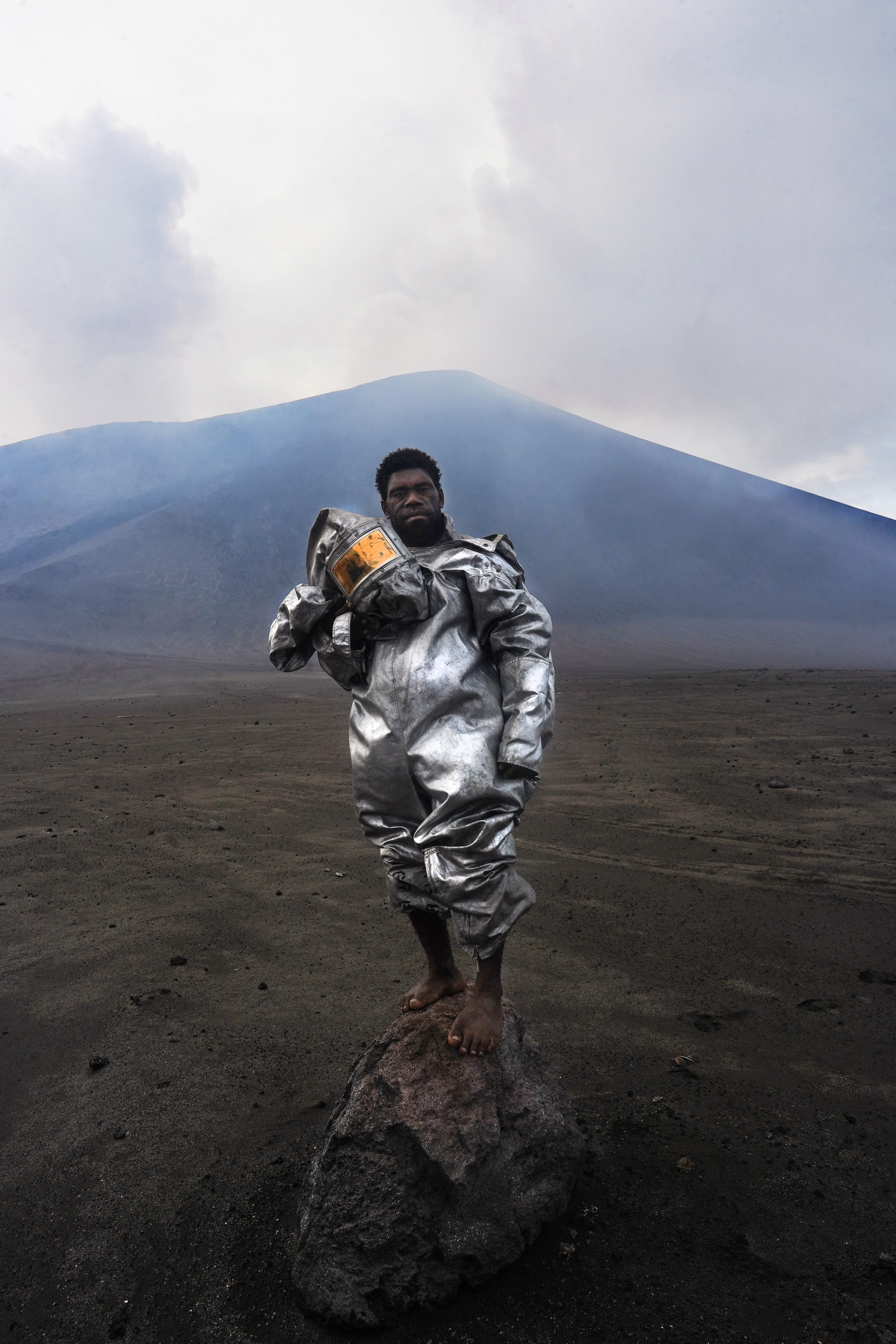 A man in a protective suit who is a volcanologist is standing barefoot atop a volcanic rock bomb on the ash plains of Mount Yasur, Vanuatu.